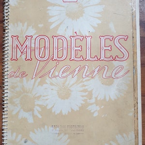 Pode incluir: Um livreto vintage de cor bege com uma coroa vermelha e o texto "MODÈLES de Vienne" em vermelho. O livreto tem uma encadernação em espiral branca e o texto "ÉTÉ 1956" em vermelho na parte inferior.