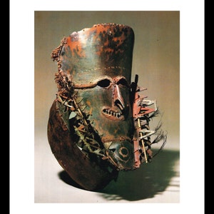 Op de afbeelding: Een houten masker met een menselijk gezicht, geschilderd in tinten groen, bruin en rood. Het masker heeft een grote, open mond met scherpe tanden en een prominente neus. Het masker is versierd met veren en andere materialen.