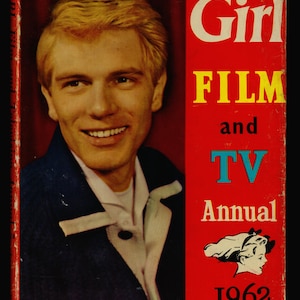 以下が含まれることがあります： 「ガール・フィルム・アンド・テレビ・アニュアル 1962」というタイトルの赤い本の表紙。表紙には、青いジャケットと白いシャツを着た金髪の若い男性が写っている。右下には、髪にリボンをつけた女の子の小さな白黒のイラストがある。