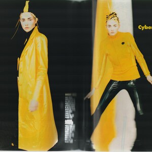 May include: Two women wearing yellow clothing. The woman on the left is wearing a long yellow coat and a black turtleneck. The woman on the right is wearing a yellow turtleneck and black pants. The text "Cyber Yellow" is printed in black on a yellow background.