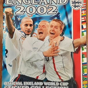 May include: A sticker collection cover featuring three soccer players in white jerseys with red accents. The cover is red, white, and blue with the text "MERLIN'S ENGLAND 2002" and "OFFICIAL ENGLAND WORLD CUP STICKER COLLECTION".