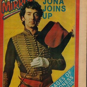 May include: A magazine cover featuring a man in a red and gold military-style jacket holding a red and black hat. The magazine is called "Record Mirror" and the headline reads "Jona Joins Up". The cover also includes the text "8 Pages of Madness Madness" and "Your Xmas Survival Kit".