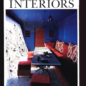 Puede incluir: La portada de la revista "The World of Interiors" presenta un diseño de interiores. La habitación tiene paredes azul oscuro, una mesa larga y asientos rojos. El título de la revista está en letras grandes y en negrita.