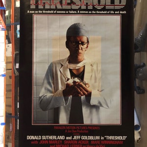 Pode incluir: Um cartaz de filme para o filme "Threshold" estrelado por Donald Sutherland e Jeff Goldblum. O cartaz apresenta uma imagem em preto e branco de um homem com um jaleco branco de médico e um gorro cirúrgico azul, segurando um instrumento médico. O texto no cartaz diz: "Um homem no limiar do sucesso ou do fracasso. Uma mulher no limiar da vida e da morte."