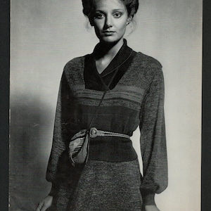 May include: A woman wearing a grey and black striped sweater with a brown leather strap and a silver buckle. The sweater has a v-neck and long sleeves.