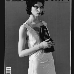 Puede incluir: Portada de revista en blanco y negro con una mujer que lleva un vestido blanco y sostiene un zapato negro. La revista se titula "RE-FOTION" y el texto "Lily McMenamy lleva Balenciaga" está en la parte inferior de la portada.