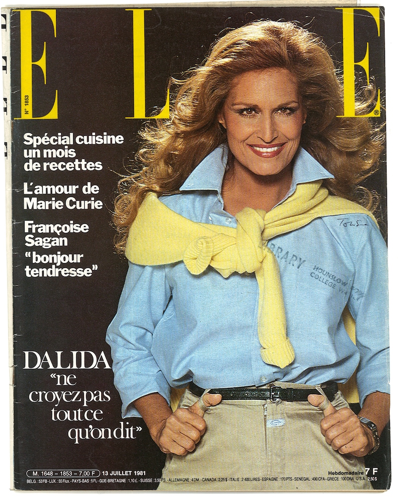 May include: Magazine cover with a woman in a light blue shirt, yellow sweater, and khaki trousers. The magazine is "ELLE", with large yellow lettering. Text includes "DALIDA" and a French phrase. The cover also features text about recipes, Marie Curie, and Fran&ccedil;oise Sagan.