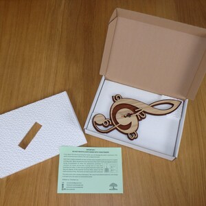 May include: A wooden clock shaped like a treble clef, packaged in a brown cardboard box with white foam padding. The clock is made of light and dark wood, with a natural wood grain finish. The clock face is visible in the centre of the treble clef, with black hands.