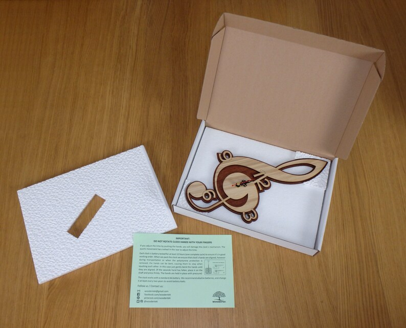 May include: A wooden clock shaped like a treble clef, packaged in a brown cardboard box with white foam padding. The clock is made of light and dark wood, with a natural wood grain finish. The clock face is visible in the centre of the treble clef, with black hands.