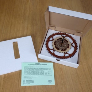 Pode incluir: Um rel&oacute;gio de madeira com design de engrenagem, embalado em uma caixa branca com um inserto de espuma. O rel&oacute;gio tem duas engrenagens, uma marrom e outra de madeira natural. Os ponteiros do rel&oacute;gio s&atilde;o pretos. A caixa inclui um cart&atilde;o de instru&ccedil;&otilde;es verde e branco com o texto "N&Atilde;O GIRE AS AGULHAS DO REL&Oacute;GIO COM OS DEDOS".