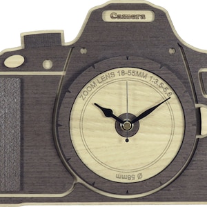Puede incluir: Un reloj de madera con forma de cámara con un acabado de madera de grano marrón. La esfera del reloj es de madera clara con manecillas negras y el texto "ZOOM LENS 18-55MM 1:3.5-5.6" y "58mm". La parte superior del reloj tiene la palabra "Camera" grabada.