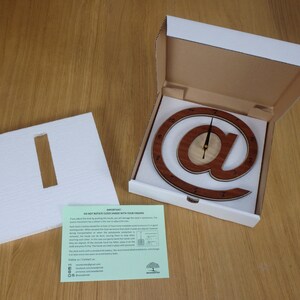 May include: A wooden clock shaped like the "@" symbol, with a white background and black hands. The clock is in a white box with a white foam insert. The box also includes a green card with instructions and contact information.