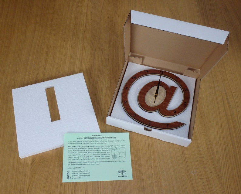 May include: A wooden clock shaped like the "@" symbol, with a white background and black hands. The clock is in a white box with a white foam insert. The box also includes a green card with instructions and contact information.