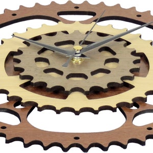 Pode incluir: Um rel&oacute;gio de parede de madeira com um anel exterior castanho e um anel interior dourado. O mostrador do rel&oacute;gio &eacute; composto por tr&ecirc;s engrenagens interligadas. Os ponteiros do rel&oacute;gio s&atilde;o pretos.