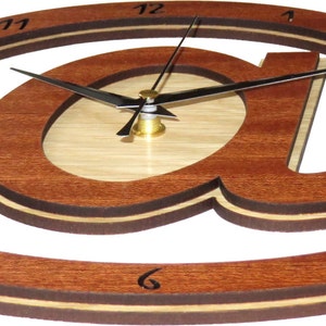 May include: A wooden wall clock shaped like the "@" symbol. The clock has a light brown wood face with a darker brown wood frame. The clock hands are black and the numbers are black on a white background.