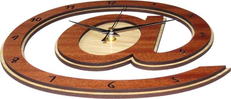 May include: A wooden wall clock shaped like the "@" symbol. The clock has a light brown wood face with a darker brown wood frame. The clock hands are black and the numbers are black on a white background.