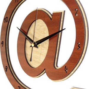 May include: A wooden wall clock with a brown and natural wood finish. The clock face is shaped like the "@" symbol. The clock hands are black and the numbers are black on a white background.