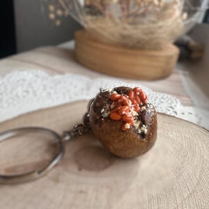 May include: A brown and white baked potato keyring with red beans and cheese on top. The keyring is attached to a silver key ring.