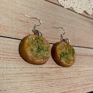 May include: A pair of dangle earrings featuring a round, brown and yellow clay charm that resembles a piece of bread with melted butter and green herbs on top.