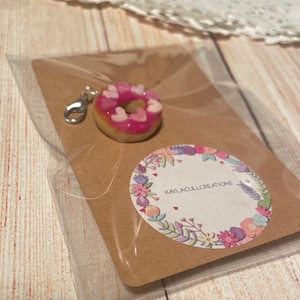 May include: A pink and white donut charm with a silver clasp. The donut is decorated with pink frosting and white hearts. The charm is packaged on a brown card with the text "KAYLACULLCREATIONS" in a white circle with floral designs.