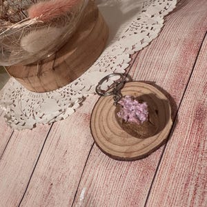 May include: A novelty keychain shaped like a baked potato with a pink filling, presented on a small wooden disc. The keychain has a silver metal ring. The background shows a wooden surface and a decorative glass dome.