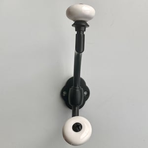 Op de afbeelding: Een zwarte metalen wandhaak met witte keramische knoppen. De haak heeft een gebogen ontwerp en een decoratieve basis. De knoppen zijn rond en hebben een zwarte schroef in het midden.