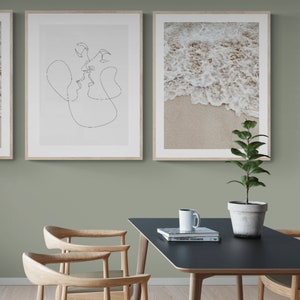 May include: Two framed prints on a sage green wall, one with a line drawing of a couple kissing, the other with a photo of ocean waves crashing on a sandy beach. A black table with a white coffee mug and a book titled "100 Graphic Designers" sits in front of the wall. A potted plant with green leaves sits on the table.