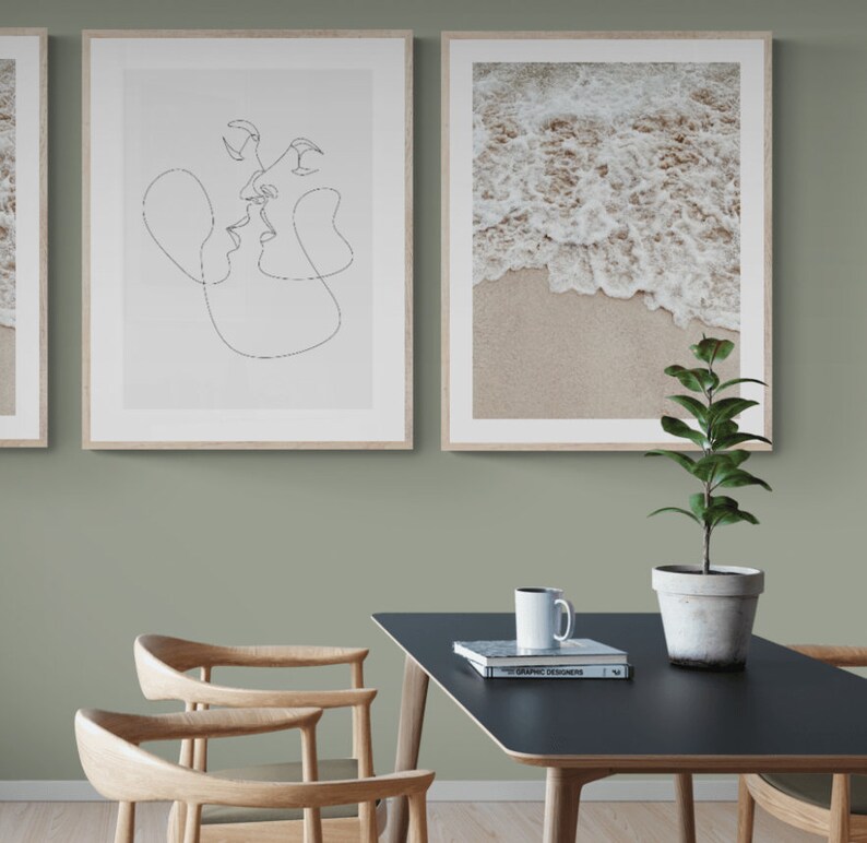May include: Two framed prints on a sage green wall, one with a line drawing of a couple kissing, the other with a photo of ocean waves crashing on a sandy beach. A black table with a white coffee mug and a book titled "100 Graphic Designers" sits in front of the wall. A potted plant with green leaves sits on the table.