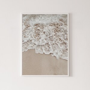 May include: A close-up of foamy white ocean waves crashing on a sandy beach. The sand is a light beige color.