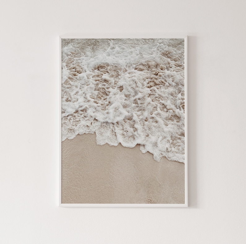 May include: A close-up of foamy white ocean waves crashing on a sandy beach. The sand is a light beige color.