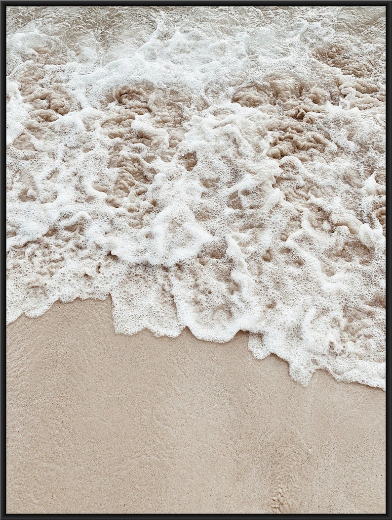 May include: Close-up of foamy white ocean waves crashing on a sandy beach. The sand is a light beige color.