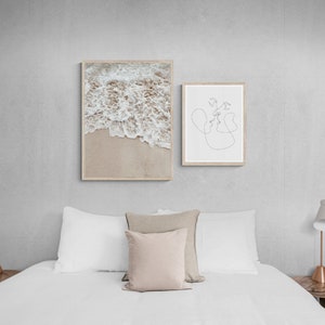 May include: Two framed prints on a white wall above a white bed with white bedding and two bedside tables with lamps. The print on the left is a photograph of ocean waves crashing on a sandy beach. The print on the right is a line drawing of two people kissing.