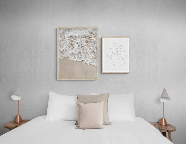 May include: Two framed prints on a white wall above a white bed with white bedding and two bedside tables with lamps. The print on the left is a photograph of ocean waves crashing on a sandy beach. The print on the right is a line drawing of two people kissing.