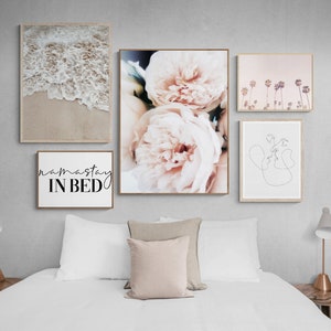 May include: Five framed prints on a white wall above a white bed with a beige headboard. The prints are a beach scene with waves, a close-up of pink peonies, a line drawing of two faces, a pink sunset with palm trees, and a white print with the text "namaste in bed".