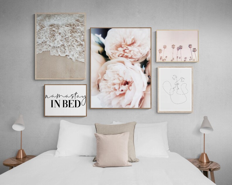 May include: Five framed prints on a white wall above a white bed with a beige headboard. The prints are a beach scene with waves, a close-up of pink peonies, a line drawing of two faces, a pink sunset with palm trees, and a white print with the text "namaste in bed".