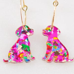 May include: A pair of pink dog-shaped earrings with gold-colored hoops. The dogs are filled with colorful, iridescent confetti-like pieces. The earrings are displayed on a white marble surface.