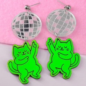 May include: A pair of novelty earrings featuring a disco ball design with a green cat charm. The cat is in a dancing pose with its arm raised. The earrings have a silver-coloured post.