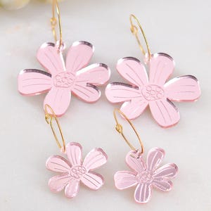 May include: Four pairs of pink daisy flower earrings with gold-colored hoops. The earrings are made of a reflective material and have a detailed center. The earrings are displayed on a white marble surface.