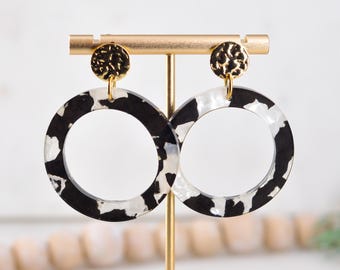 May include: A pair of black and white patterned hoop earrings with gold textured accents. The earrings are made of acrylic and have a modern, geometric design.