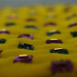 May include: Close-up of a yellow fabric with a textured surface. The fabric is adorned with small, rectangular embellishments in teal and purple, each accented with a gold bar. The overall effect is a pattern of raised elements.