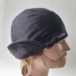 Könnte beinhalten: Eine einfarbige schwarze Kappe mit strukturierter Krone und umgeschlagenem Rand. Der Stoff weist sichtbare Nähte auf und ein kleines gesticktes Logo an der Seite mit der Aufschrift CORAL MOON creations in rosa und weißem Garn über dem Rand.