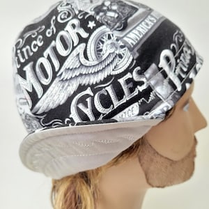 May include: A black and white motorcycle-themed skull cap featuring graphic prints of wings, wheels, and the text "Prince of MOTOR CYCLES". The cap has a solid light gray brim with visible stitching and a rounded crown design made of patterned fabric.