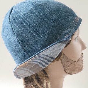 May include: A blue denim cap with a rounded crown and a short, turned-up brim. The underside of the brim features a grey and brown plaid pattern with horizontal stripes. The cap is constructed from multiple panels of fabric stitched together.