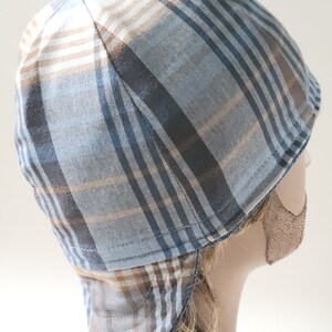 May include: A welding cap featuring a plaid pattern in shades of light blue, dark blue, white, and tan. The cap is constructed with multiple fabric panels and includes a protective neck flap at the back, stitched with visible thread along the seams.