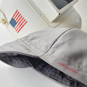 May include: A light gray welding cap featuring a dark gray quilted interior lining and the text CORAL MOON embroidered in red thread on the side. The cap is positioned in front of a white welding helmet that displays a small American flag decal.
