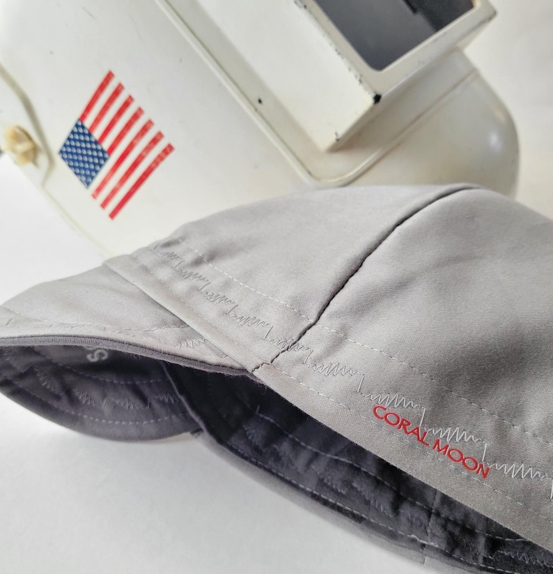 May include: A light gray welding cap featuring a dark gray quilted interior lining and the text CORAL MOON embroidered in red thread on the side. The cap is positioned in front of a white welding helmet that displays a small American flag decal.