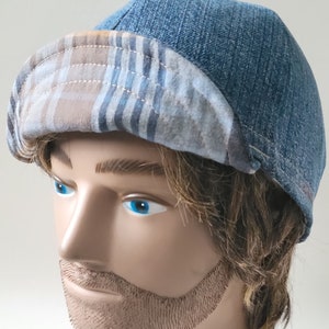 May include: A blue denim cap featuring a rounded crown and a folded brim. The underside of the brim displays a tan, blue, and white plaid pattern. The cap is constructed with visible stitching along the seams and the brim edge, creating a structured look.
