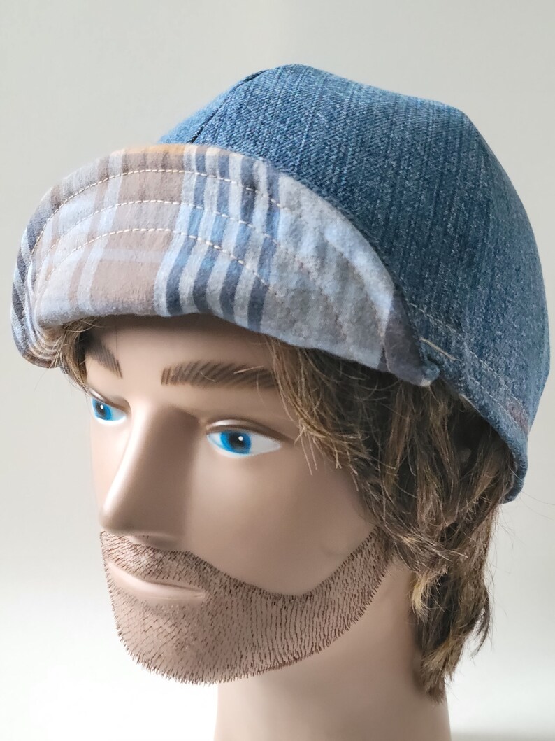 May include: A blue denim cap featuring a rounded crown and a folded brim. The underside of the brim displays a tan, blue, and white plaid pattern. The cap is constructed with visible stitching along the seams and the brim edge, creating a structured look.