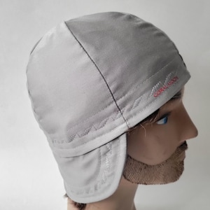 May include: A light gray cap with ear flaps and visible stitching along the seams. The text CORAL MOON is embroidered in red thread on the side of the cap. The cap features a multi-panel construction with a rounded crown and a structured brim.