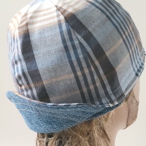 May include: A brimless cap featuring a plaid pattern in shades of blue, grey, and tan. The hat has a structured crown with a folded, denim-textured blue band around the base, showcasing visible stitching along the edge of the folded fabric.